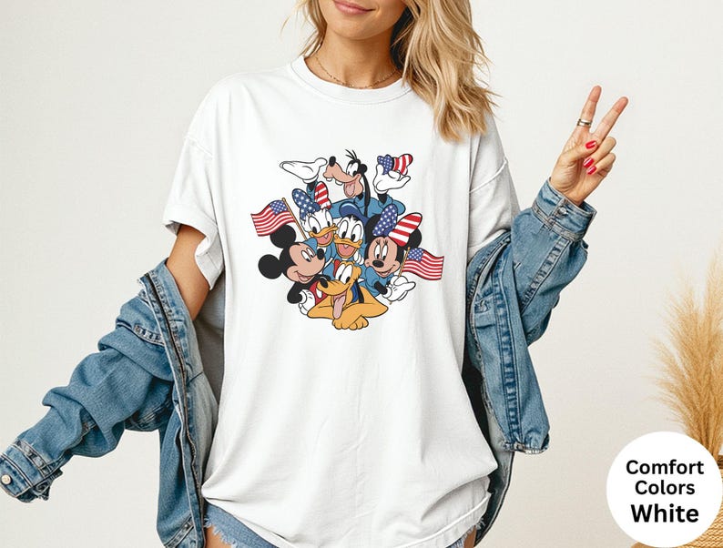 May include: White Comfort Colors t-shirt featuring a graphic of Mickey Mouse, Minnie Mouse, Donald Duck, Goofy, and Pluto, each holding or wearing patriotic American flags. The shirt has a crew neck and short sleeves.