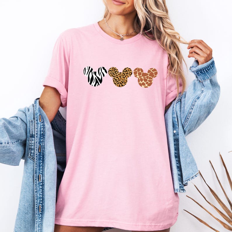 May include: Pink t-shirt featuring three Mickey Mouse head silhouettes with animal print designs: zebra, leopard, and giraffe. The shirt has short sleeves and a crew neck. A denim jacket is draped over the arm.
