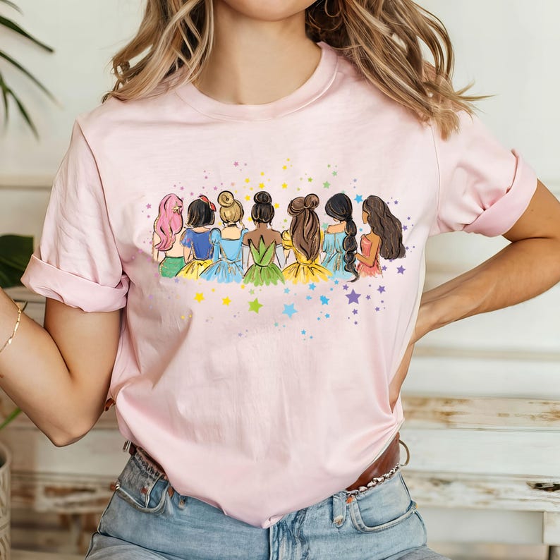 May include: Pink t-shirt with a graphic of seven princesses from Disney's animated films. The princesses are standing in a line, facing away from the viewer. They are wearing their signature dresses and have their hair styled in their signature ways. The graphic is surrounded by a scattering of stars.