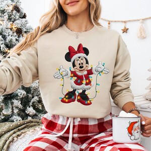 May include: Beige crew neck sweatshirt with a festive Minnie Mouse design. Minnie is in a red Santa-style outfit with a matching bow and shoes, holding Christmas lights. The sweatshirt is paired with red and white plaid pyjama bottoms.