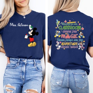Puede incluir: Una camiseta azul marino con un gr&aacute;fico de Mickey Mouse en la parte delantera y el texto "Mrs. Alisson" en el lado izquierdo del pecho. La parte posterior de la camiseta tiene un gr&aacute;fico colorido con el texto "&iexcl;Bienvenido a nuestra aula donde la magia nunca se detiene y la aventura nunca termina!"