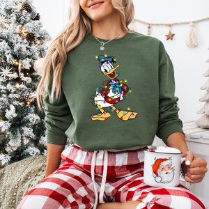 K&ouml;nnte beinhalten: Gr&uuml;nes Sweatshirt mit einem Cartoon-Design von Donald Duck, umwickelt mit Weihnachtsbeleuchtung und h&auml;lt ein Geschenk. Dazu eine rot-wei&szlig; karierte Pyjamahose. Eine Santa-Tasse wird in der Hand gehalten.