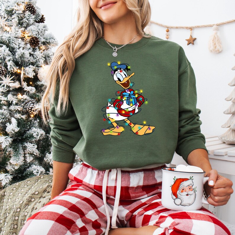 K&ouml;nnte beinhalten: Gr&uuml;nes Sweatshirt mit einem Cartoon-Design von Donald Duck, umwickelt mit Weihnachtsbeleuchtung und h&auml;lt ein Geschenk. Dazu eine rot-wei&szlig; karierte Pyjamahose. Eine Santa-Tasse wird in der Hand gehalten.