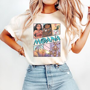 May include: A white t-shirt with a cartoon graphic of characters from the Disney movie Moana. The graphic includes Moana, Maui, Pua the pig, Hei Hei the rooster, and the Kakamora.