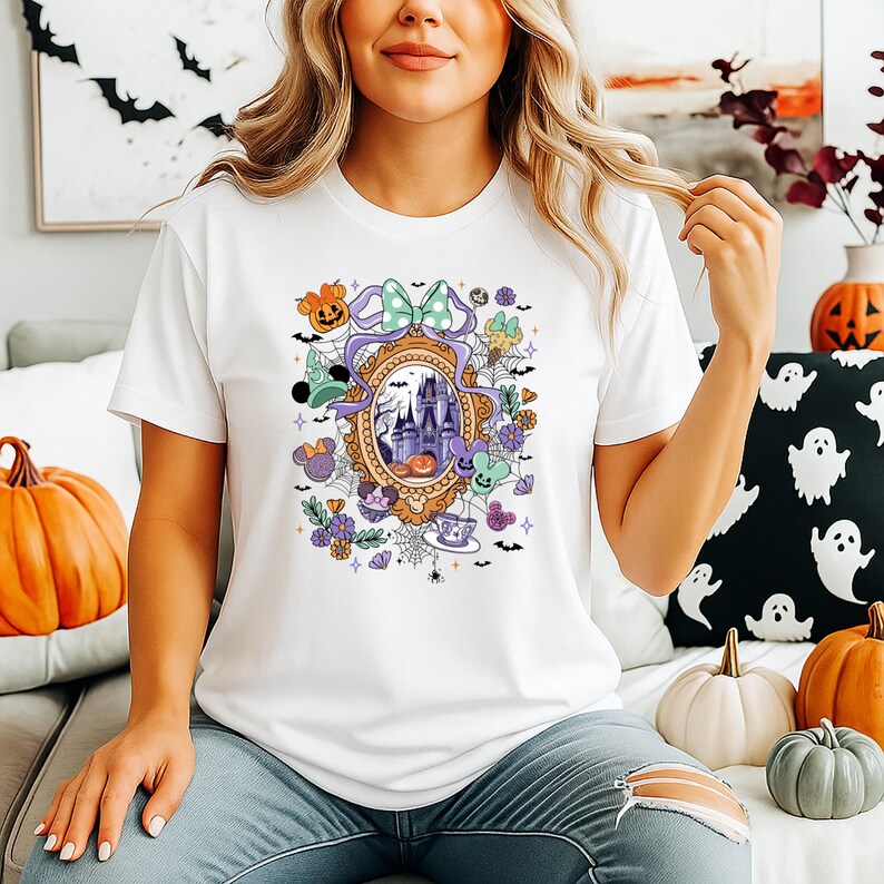 Puede incluir: Camiseta blanca con un dise&ntilde;o tem&aacute;tico de Halloween. El gr&aacute;fico incluye un castillo, calabazas y otros elementos festivos dentro de un marco decorativo. Un lazo verde menta y detalles morados completan el dise&ntilde;o.
