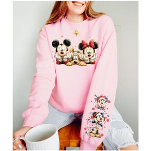 May include: Pink sweatshirt featuring Mickey Mouse, Minnie Mouse, and Pluto in a Christmas design. The sleeve has additional Christmas-themed character graphics. A gold star and other small stars are also on the sweatshirt.