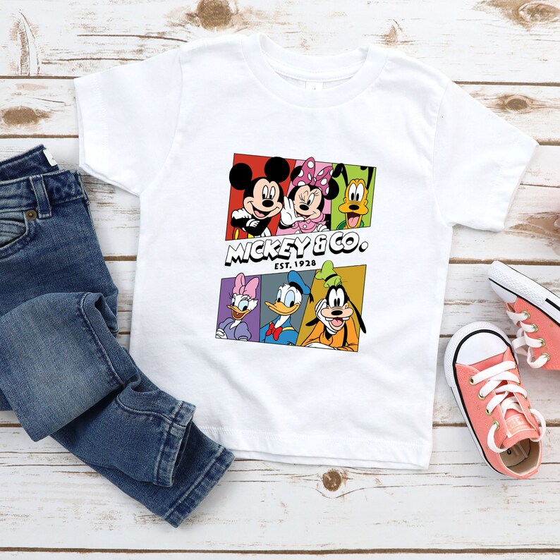 Pu&ograve; includere: Maglietta bianca con una grafica colorata di Topolino, Minnie Mouse, Paperino, Pippo e Pluto. La grafica include il testo "Mickey & Co. Est. 1928".