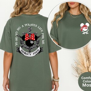 May include: Moss green t-shirt featuring a pirate-themed design. The back of the shirt displays an anchor with a Minnie Mouse head, a red polka-dot bow, and the text "Yo Ho Ho A Pirates Life For Me Disney 2026". The front has a smaller pirate Minnie Mouse design.