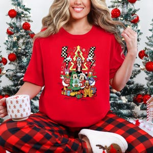 May include: Red t-shirt featuring a Christmas-themed graphic with cartoon characters. The design includes Mickey Mouse, Minnie Mouse, Donald Duck, and Goofy, with a Christmas tree and lightning bolt accents. The person is wearing red and black plaid pajama pants.
