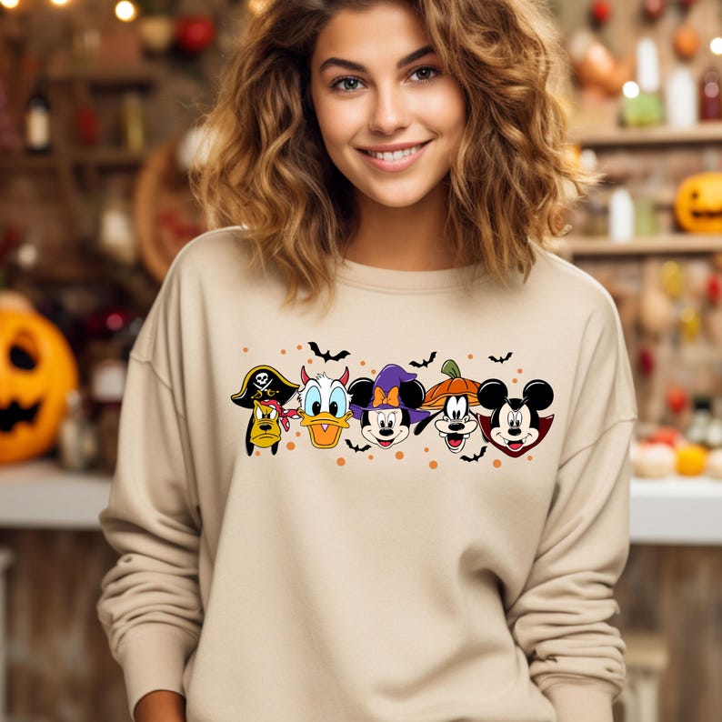 Op de afbeelding: Een beige sweatshirt met een Halloween-thema ontwerp met Mickey Mouse, Donald Duck, Goofy en Pluto in kostuums. De personages staan in een rij met vleermuizen en oranje stippen op de achtergrond.