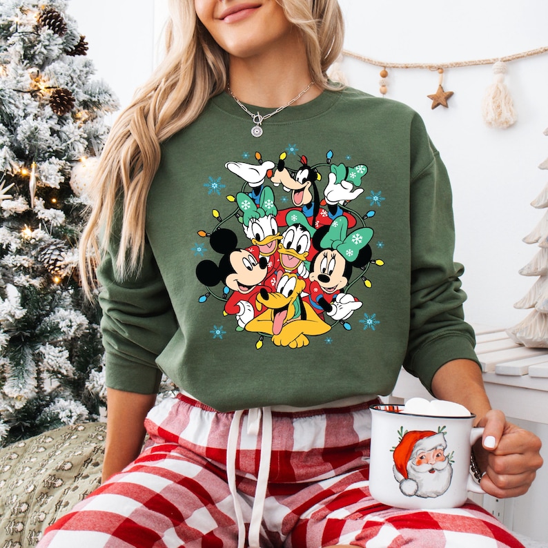 K&ouml;nnte beinhalten: Gr&uuml;nes Sweatshirt mit einem festlichen Design von Disney-Figuren, darunter Mickey Mouse, Minnie Mouse, Donald Duck und Goofy, umgeben von Lichterketten und Schneeflocken. Die Person tr&auml;gt eine rot-wei&szlig; karierte Pyjamahose und h&auml;lt eine Weihnachtsmann-Tasse.