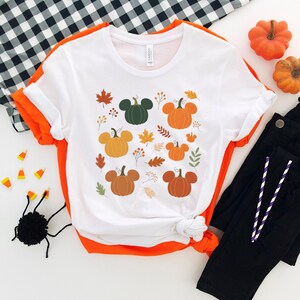 May include: White t-shirt with a fall-themed design featuring pumpkins with mouse ears, autumn leaves, and sprigs. The shirt is folded with orange trim and paired with black pants. Decorative pumpkins and a spider are also visible.