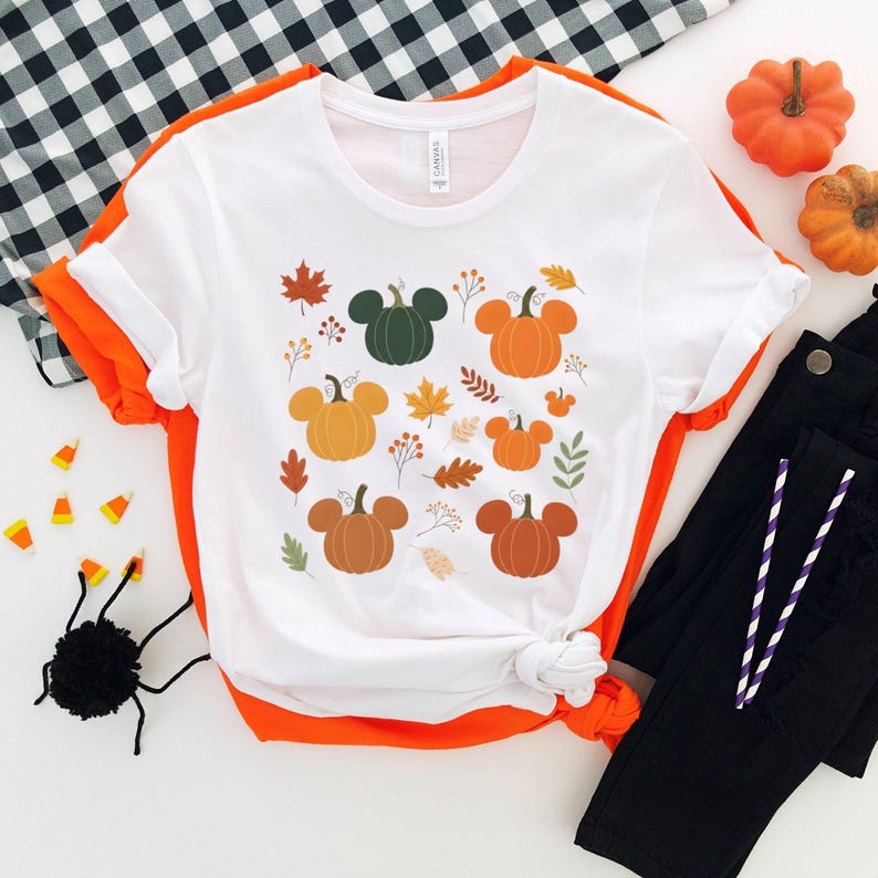 May include: White t-shirt with a fall-themed design featuring pumpkins with mouse ears, autumn leaves, and sprigs. The shirt is folded with orange trim and paired with black pants. Decorative pumpkins and a spider are also visible.