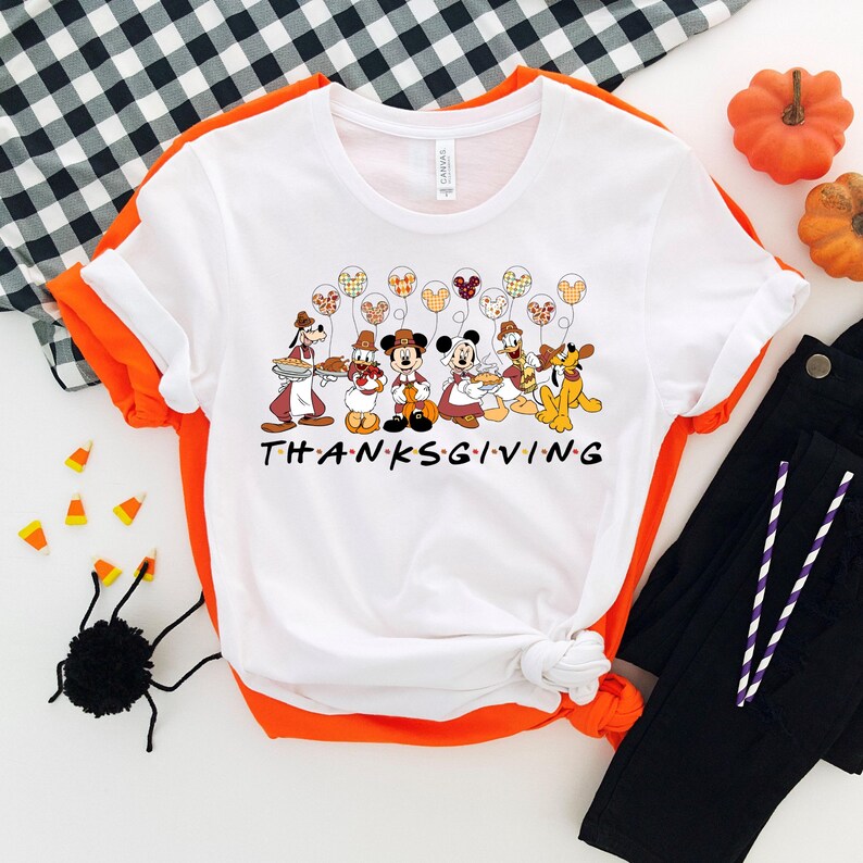 K&ouml;nnte beinhalten: Wei&szlig;es T-Shirt mit Thanksgiving-Motiv, auf dem Mickey Mouse und seine Freunde Teller mit Essen halten. Die Figuren sind von Ballons mit Thanksgiving-Muster umgeben. Der Text "THANKSGIVING" ist unter den Figuren gedruckt.