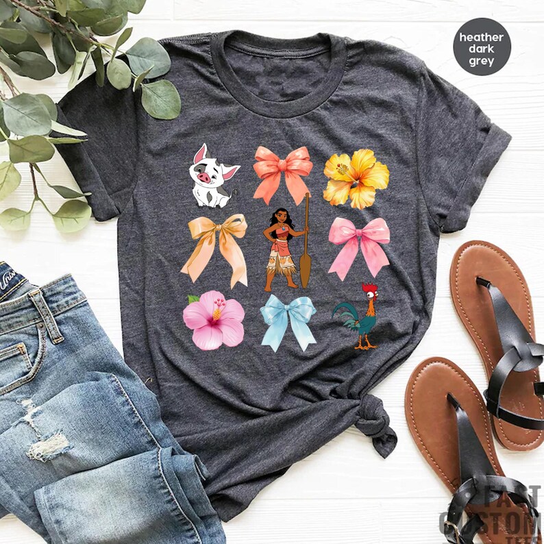 Pu&ograve; includere: Una maglietta color grigio scuro melange con una variet&agrave; di immagini colorate stampate su di essa. Le immagini includono un maiale, un fiore, un fiocco, una donna con una pagaia e un gallo.