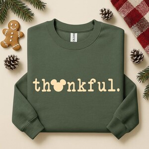 Puede incluir: Una sudadera verde oliva con la palabra "thankful" en color crema. La "o" de la palabra se reemplaza con una cabeza de Mickey Mouse. La sudadera est&aacute; rodeada de pi&ntilde;as, un hombre de jengibre y una manta a cuadros rojos y blancos.