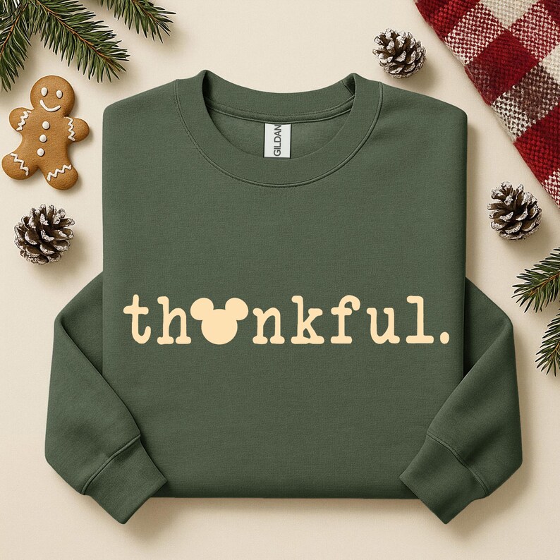 Puede incluir: Una sudadera verde oliva con la palabra "thankful" en color crema. La "o" de la palabra se reemplaza con una cabeza de Mickey Mouse. La sudadera est&aacute; rodeada de pi&ntilde;as, un hombre de jengibre y una manta a cuadros rojos y blancos.