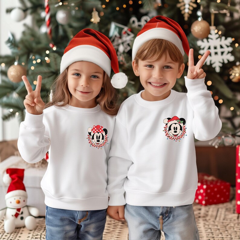 May include: Two children wearing white Christmas sweaters with a Mickey Mouse and Minnie Mouse design. The children are wearing red Santa hats and smiling.
