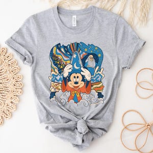 May include: Gray t-shirt with a colorful graphic of Mickey Mouse as a sorcerer. The graphic includes a blue hat, orange robe, and a swirling background with stars and clouds. The text "Mickey Mouse" is visible on the shirt.