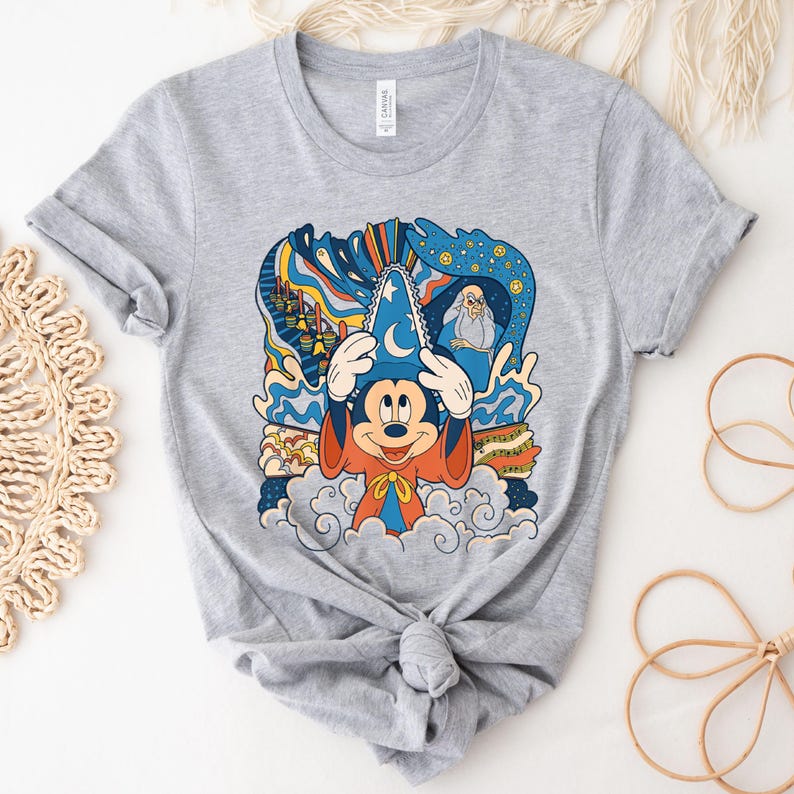 May include: Gray t-shirt with a colorful graphic of Mickey Mouse as a sorcerer. The graphic includes a blue hat, orange robe, and a swirling background with stars and clouds. The text "Mickey Mouse" is visible on the shirt.