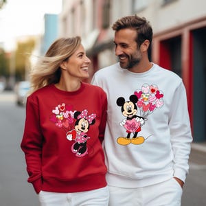 May include: A couple wearing matching red and white sweatshirts with a Mickey Mouse and Minnie Mouse design. The sweatshirts feature Mickey and Minnie holding heart-shaped balloons.