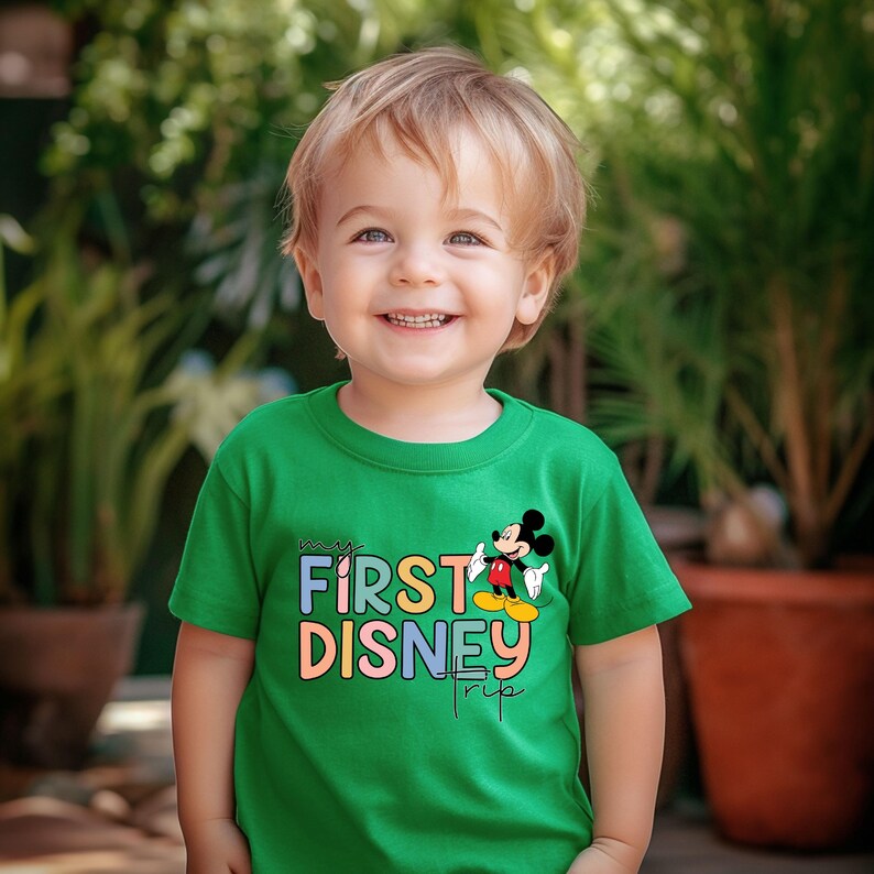 May include: A young child wearing a green t-shirt with the text "my first disney trip" in colorful letters and a cartoon image of Mickey Mouse.