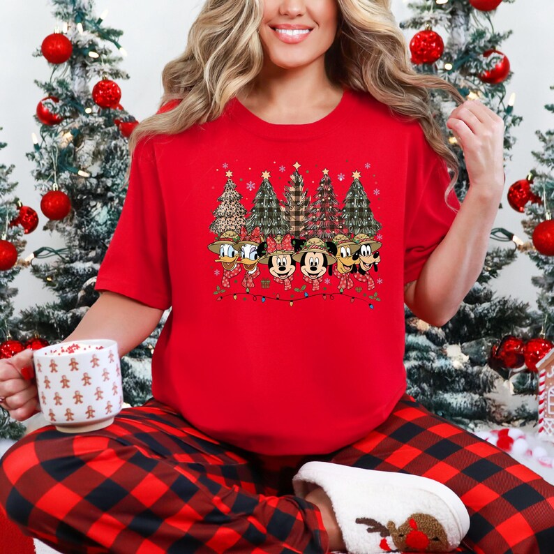 May include: A red short-sleeved t-shirt featuring a festive design. The graphic includes cartoon characters, Christmas trees, and string lights. The characters are wearing hats and scarves. The shirt is part of a holiday collection.