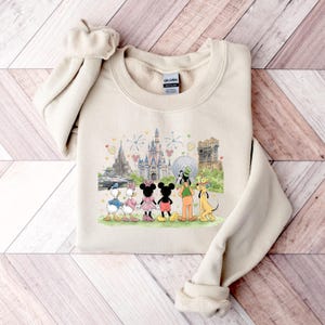May include: A beige sweatshirt featuring a colorful graphic of Disney characters, including Mickey and Minnie Mouse, Donald and Daisy Duck, Goofy, and Pluto, in front of various park landmarks. The sweatshirt is folded on a wooden surface.