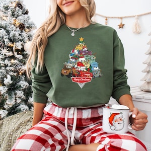May include: Green sweatshirt featuring a Christmas tree design made of cartoon cars. The tree is topped with a gold star and has a banner that reads "Merry Christmas". The sweatshirt is paired with red and white plaid pajama pants.
