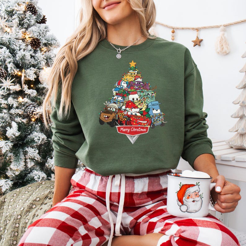 May include: Green sweatshirt featuring a Christmas tree design made of cartoon cars. The tree is topped with a gold star and has a banner that reads "Merry Christmas". The sweatshirt is paired with red and white plaid pajama pants.