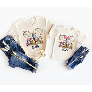 May include: Two cream colored t-shirts with a graphic of Mickey Mouse and friends in front of Cinderella's Castle. The text on the shirts reads "Disney Trip 2025".