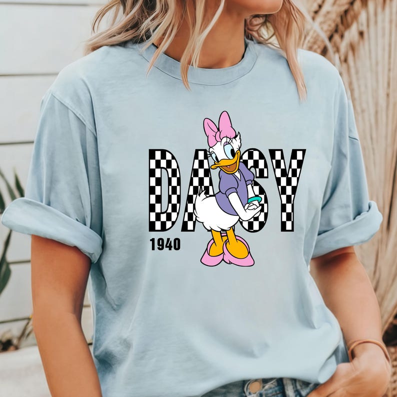May include: Light blue t-shirt with a black and white checkered graphic of the word "Daisy" with a cartoon image of Daisy Duck in the center. The year "1940" is printed below the graphic.