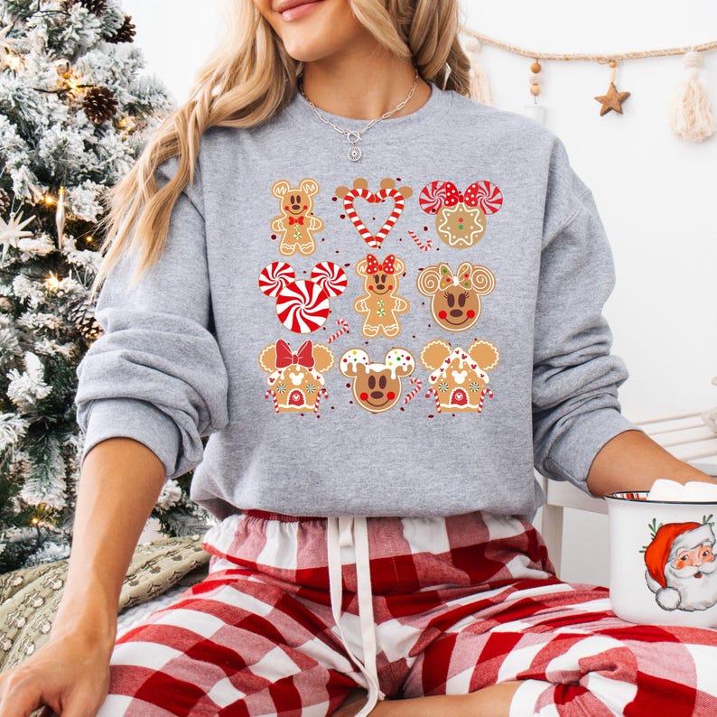 May include: A gray sweatshirt with a Christmas theme featuring gingerbread cookies shaped like Mickey Mouse and friends. The cookies are decorated with red and white icing and candy canes. The sweatshirt is worn by a person wearing red and white plaid pants.