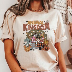 Op de afbeelding: Beige T-shirt met de tekst "ANIMAL KINGDOM" in bruine letters. De afbeelding toont stripfiguren in een truck met pompoenen. Het shirt heeft korte mouwen en een ronde hals.