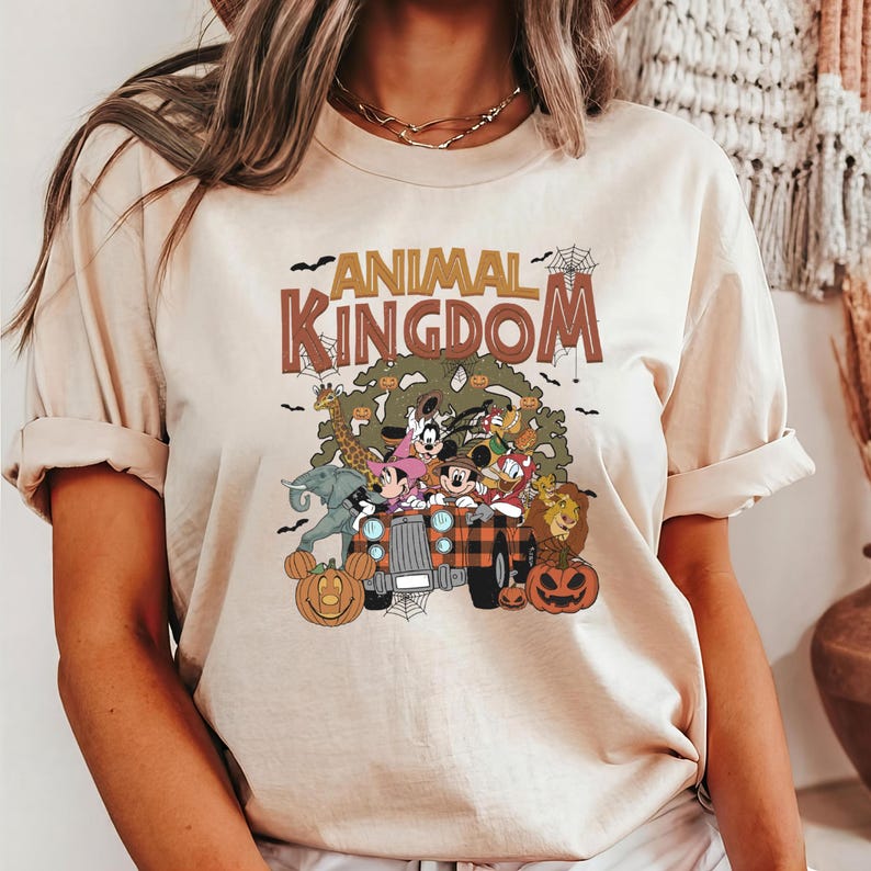 Op de afbeelding: Beige T-shirt met de tekst "ANIMAL KINGDOM" in bruine letters. De afbeelding toont stripfiguren in een truck met pompoenen. Het shirt heeft korte mouwen en een ronde hals.