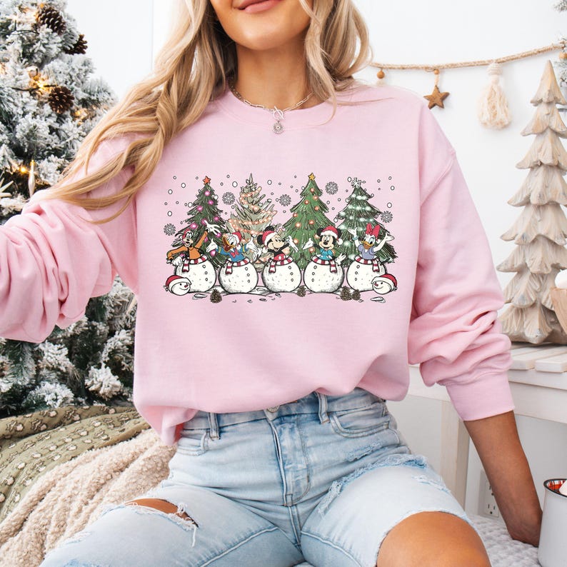 Op de afbeelding: Roze trui met een afbeelding van Disney-figuren die verkleed zijn als sneeuwpoppen voor kerstboom. De figuren zijn Mickey Mouse, Minnie Mouse, Donald Duck, Goofy en Pluto.