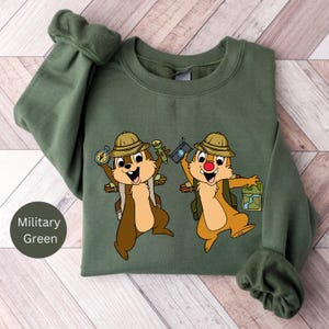 May include: A military green sweatshirt featuring a printed design of two cartoon chipmunks wearing explorer hats and carrying backpacks. The chipmunks are holding a compass, torch, camera, and map.