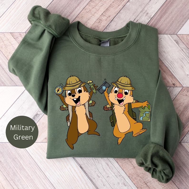 May include: A military green sweatshirt featuring a printed design of two cartoon chipmunks wearing explorer hats and carrying backpacks. The chipmunks are holding a compass, torch, camera, and map.