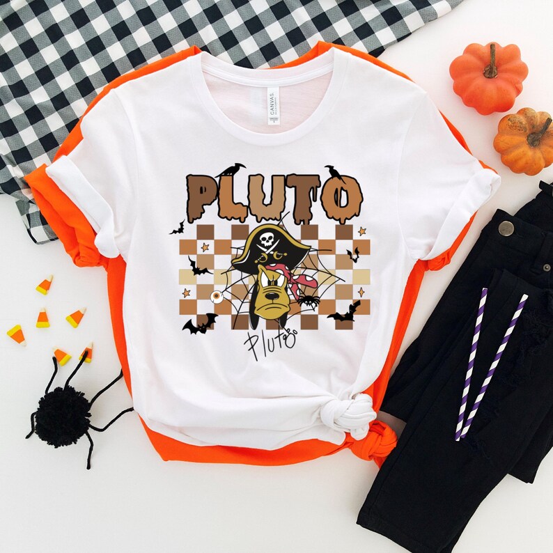 May include: White t-shirt with orange trim featuring a Halloween-themed Pluto graphic. The design includes the word "PLUTO" in brown letters, a pirate hat, spiderwebs, bats, and Pluto's face. Black pants and pumpkins are also visible.