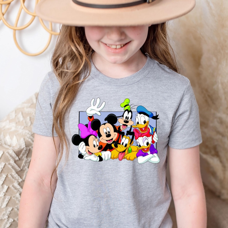 May include: A gray t-shirt featuring a colorful graphic of Disney characters, including Mickey and Minnie Mouse, Goofy, Donald Duck, and Daisy Duck. The cartoon characters are in a rectangular frame. The shirt is likely for children.