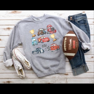 May include: A gray sweatshirt with a colorful cartoon design of cars, trophies, and checkered flags. The sweatshirt is laid out on a wooden surface with a football and jeans.