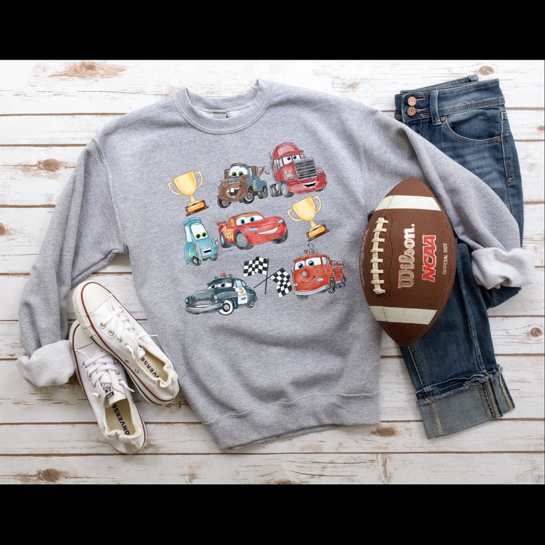 May include: A gray sweatshirt with a colorful cartoon design of cars, trophies, and checkered flags. The sweatshirt is laid out on a wooden surface with a football and jeans.