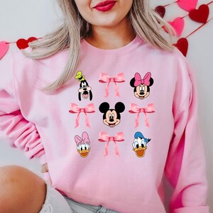 May include: Pink sweatshirt with a graphic print of Mickey Mouse, Minnie Mouse, Donald Duck, Goofy, and pink bows.