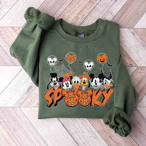 May include: Green sweatshirt with a Halloween design. The graphic features cartoon characters, Mickey Mouse balloons, bats, spiderwebs, and the word "SP00KY" in orange. The sweatshirt has long sleeves and a crew neck.