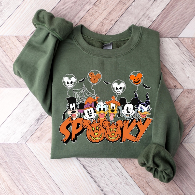May include: Green sweatshirt with a Halloween design. The graphic features cartoon characters, Mickey Mouse balloons, bats, spiderwebs, and the word "SP00KY" in orange. The sweatshirt has long sleeves and a crew neck.