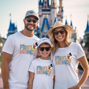 May include: A family of three wearing white t-shirts with the text "my first disney trip" in colorful letters and a cartoon image of Mickey Mouse. The shirts are a fun way to celebrate a first trip to Disney.