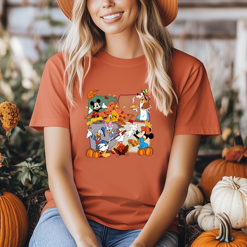 May include: Rust-colored t-shirt featuring a graphic of Disney characters celebrating Thanksgiving. Mickey Mouse, Minnie Mouse, Donald Duck, and Goofy are depicted with pumpkins and autumn leaves. The shirt is made of a soft material.