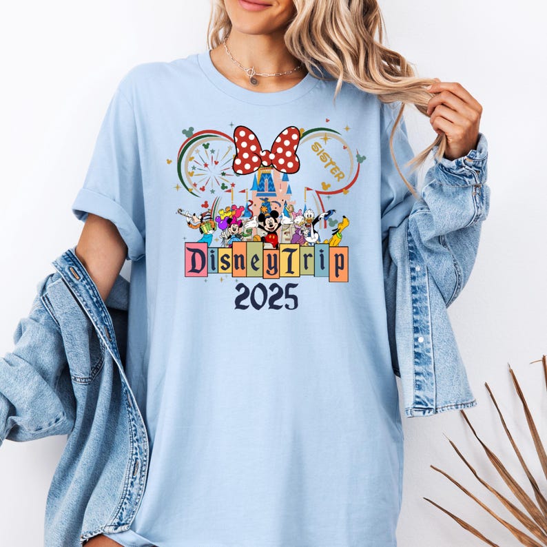 May include: Light blue t-shirt with a graphic of Mickey Mouse and friends in front of Cinderella's Castle. The text "Disney Trip 2025" is below the graphic. A red bow with white polka dots is above the castle. The text "Sister" is to the right of the bow.