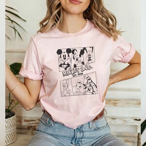 K&ouml;nnte beinhalten: Ein pinkfarbenes T-Shirt mit einem schwarz-wei&szlig;en Grafikdruck von Mickey Mouse, Minnie Mouse, Donald Duck und Goofy. Die Grafik enth&auml;lt den Text "Mickey & Co. Est. 1928".