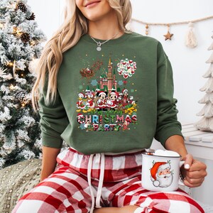 May include: Green Christmas sweatshirt featuring a colorful graphic of Disney characters, a castle, and Santa's sleigh. The text "Very Merry Christmas Party" is displayed below the characters. The person is wearing red and white plaid pajama pants and holding a mug with a Santa Claus design.